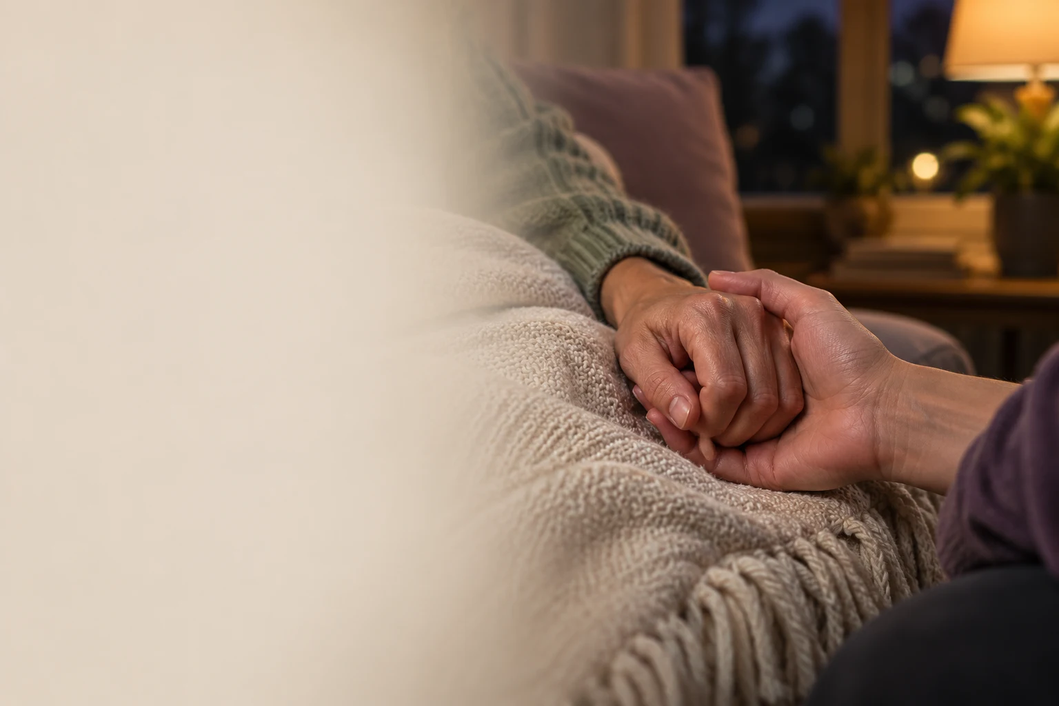 One adult’s tense hand gently supported in another’s open palm over a cream blanket in a lamplit room, with no faces visible.